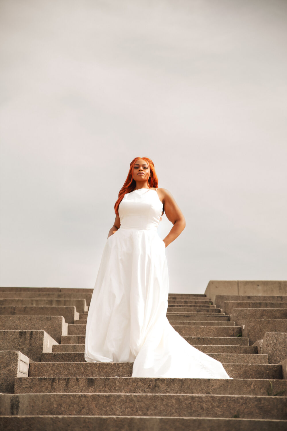 A woman in a sleeveless white wedding gown stands on a large outdoor staircase, looking forward with her hands in her pockets.