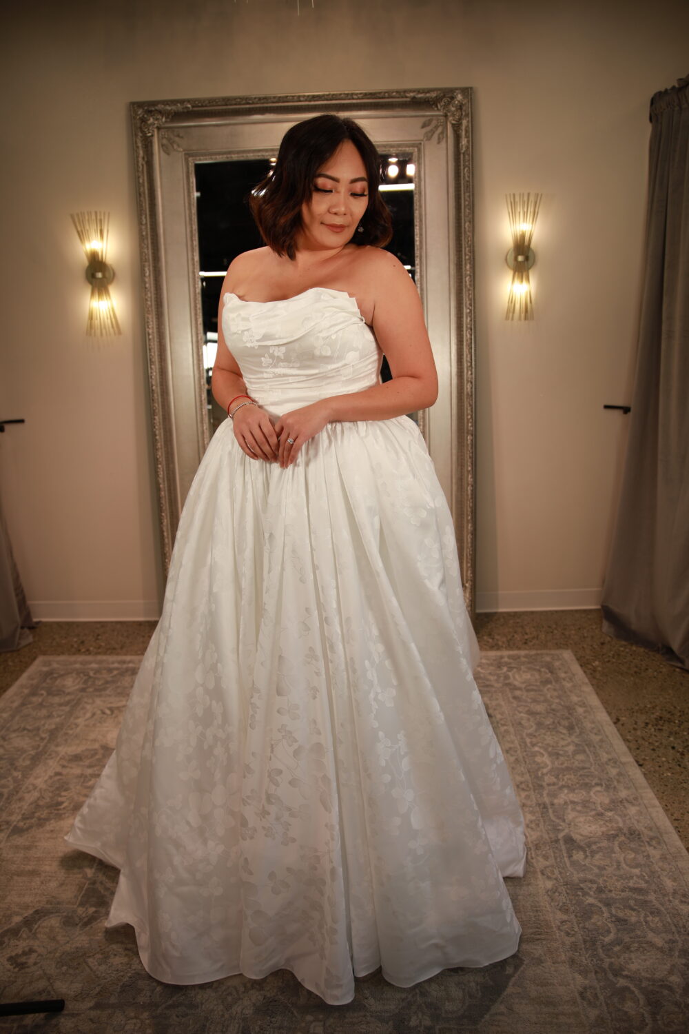 A woman in a strapless patterned ball gown poses indoors in front of a large framed mirror, gently holding the skirt of her dress.