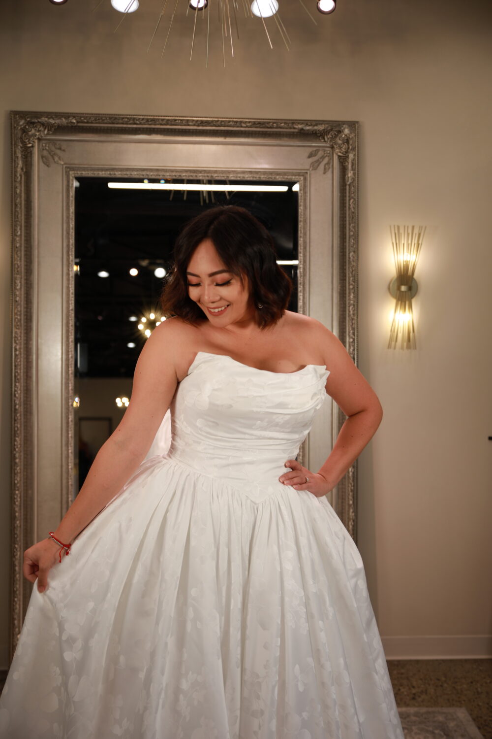 A woman in a strapless ball gown with a floral-textured fabric smiles while looking down, standing in front of a large ornate mirror indoors.