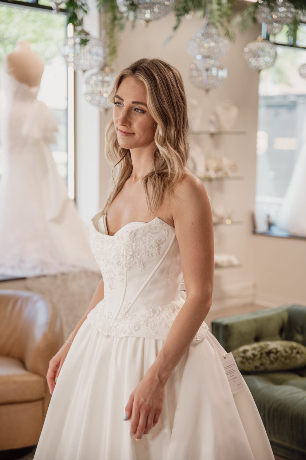 A bride in a strapless embroidered ball gown looks off to the side while standing in a bridal boutique with hanging crystal décor.