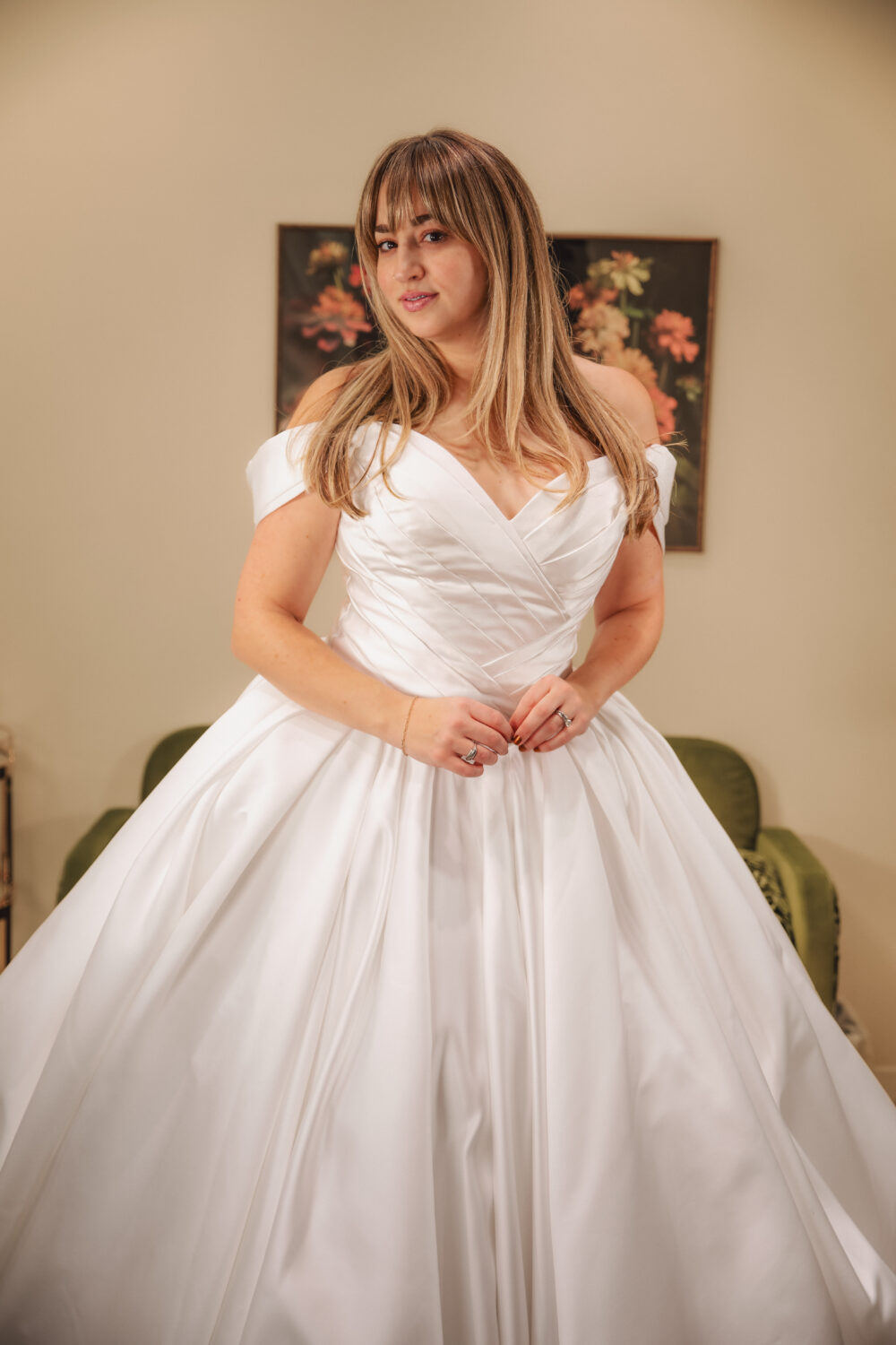 A bride in an off-the-shoulder satin ball gown looks confidently at the camera while gently holding the front of her dress.
