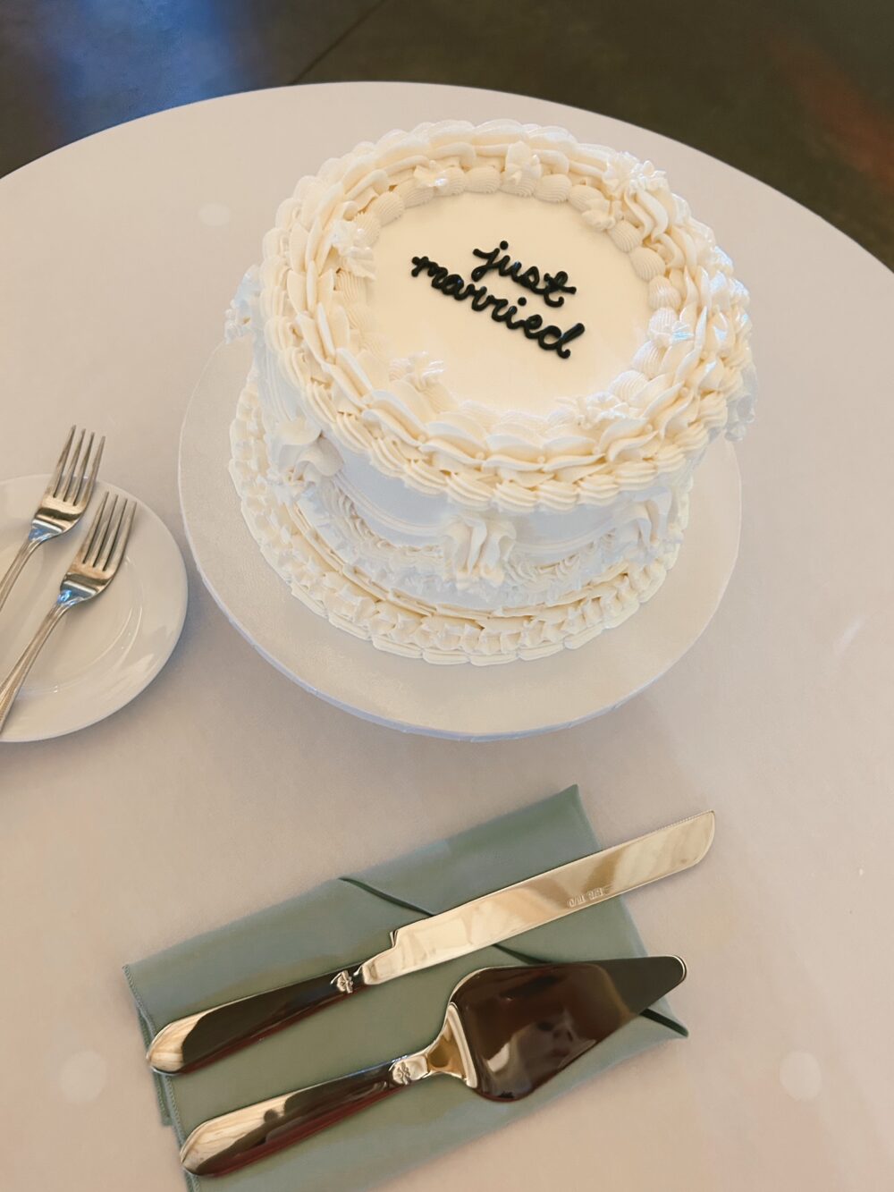 A small white wedding cake decorated with piped frosting sits on a table beside a cake knife, server, and two forks.