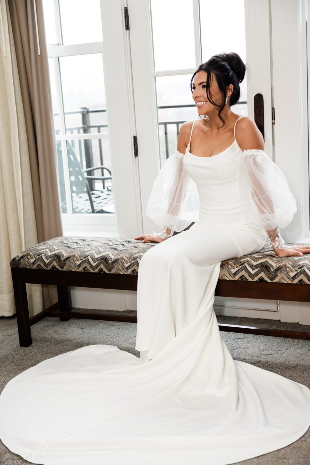 A bride in a fitted white gown with sheer puff sleeves sits on a bench near a bright window, smiling as she looks to the side.
