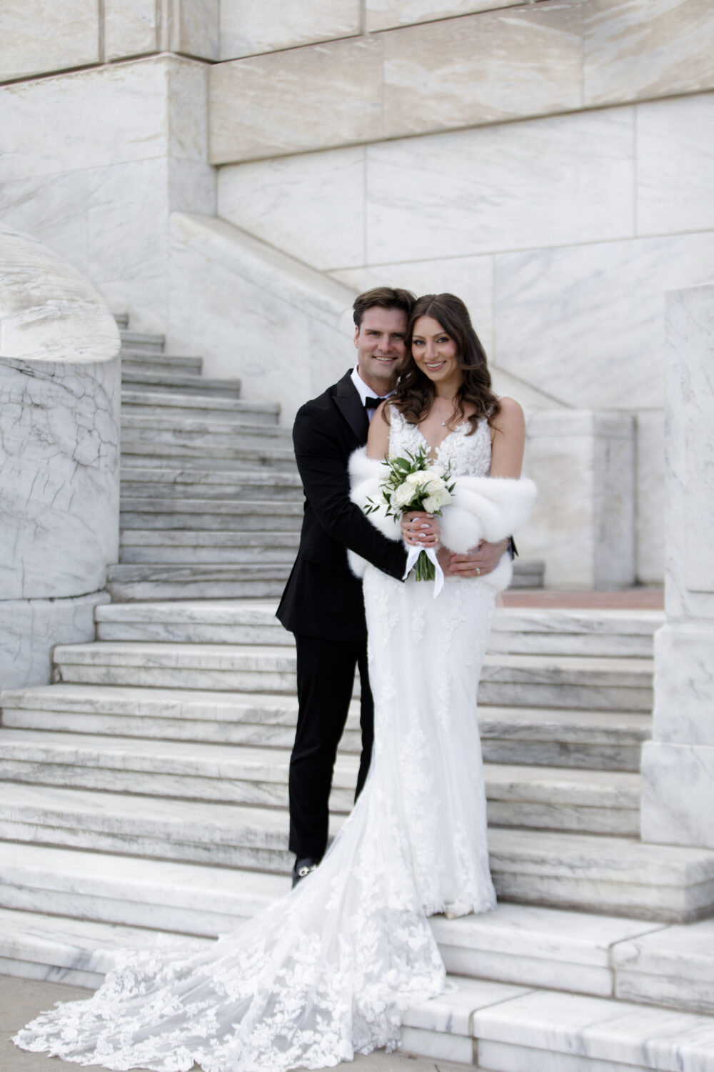 A bride and groom stand together on marble steps, with the groom hugging the bride from behind as she holds a small white bouquet.