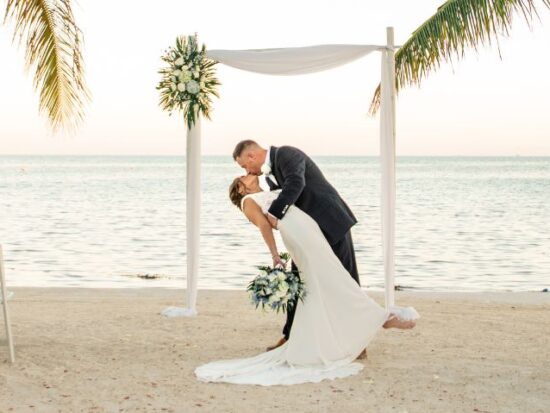 A groom dips his bride under a simple beach wedding arch as they kiss, with the ocean and palm trees in the background.