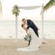 A groom dips his bride under a simple beach wedding arch as they kiss, with the ocean and palm trees in the background.