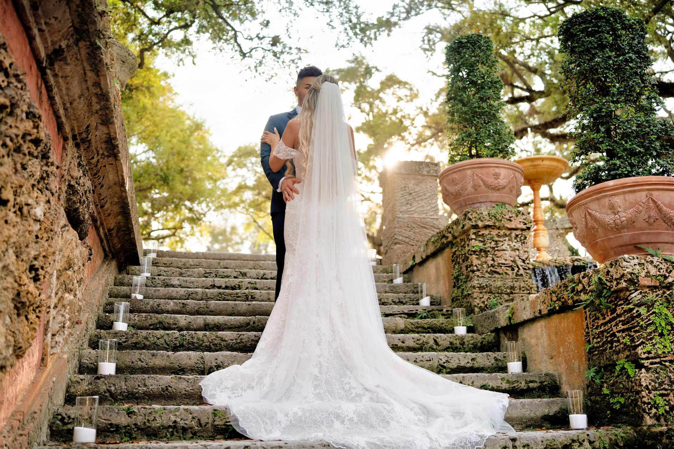 A bride in a long lace wedding gown walking up stone steps surrounded by greenery and warm-toned architecture.