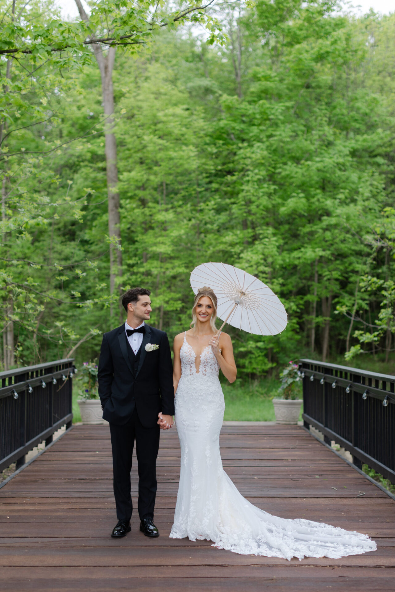 A bride and groom standing on a wooden bridge surrounded by lush spring greenery, with the bride holding a white parasol and wearing a long gown.