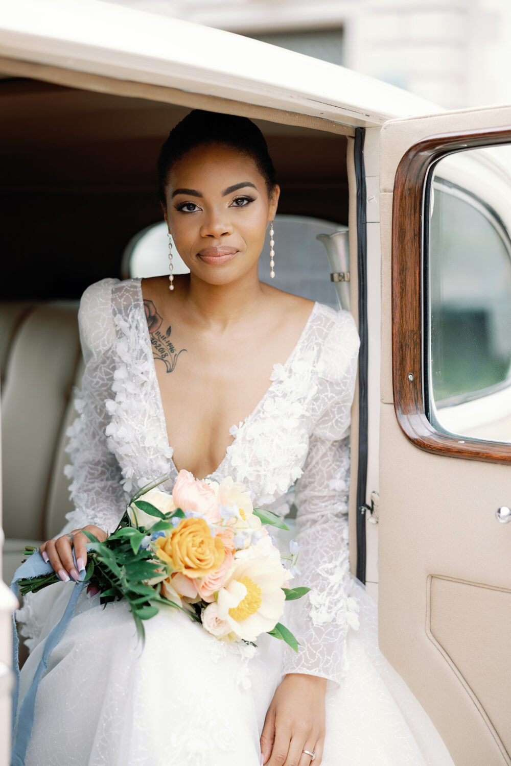 Bride in deep V-neck lace dress holding bouquet while sitting in a vintage car.