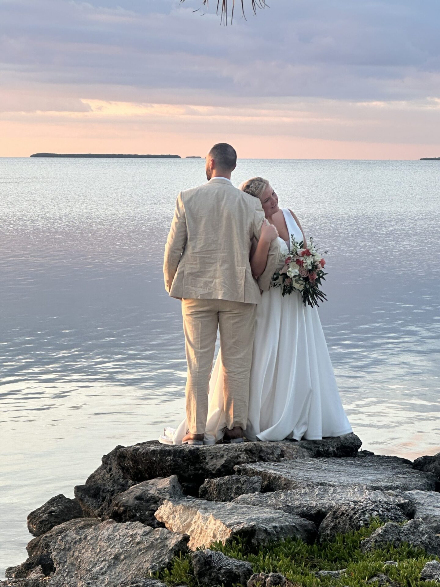 A couple standing on rocks beside a calm lake at sunset, with the bride wearing a flowing gown and holding a bouquet.
