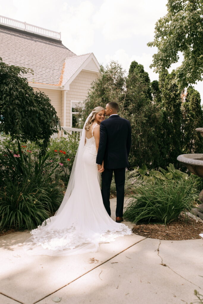 Bride and groom kissing on a garden pathway outside a house, bride wearing a long veil and fitted gown.