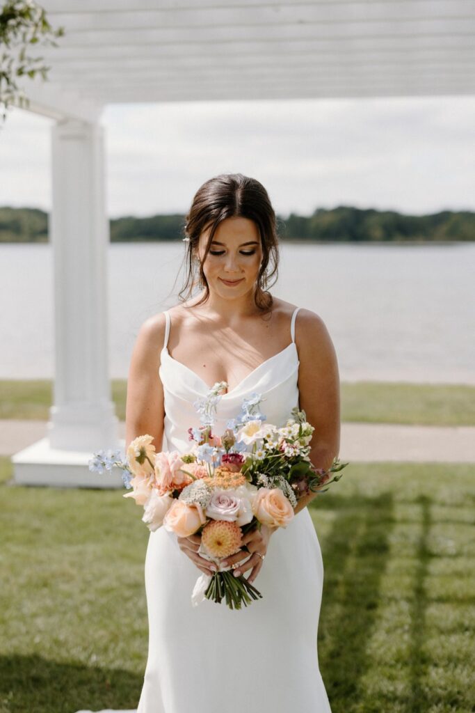Bride in a minimalist white gown holding a pastel bouquet by a lakeside.