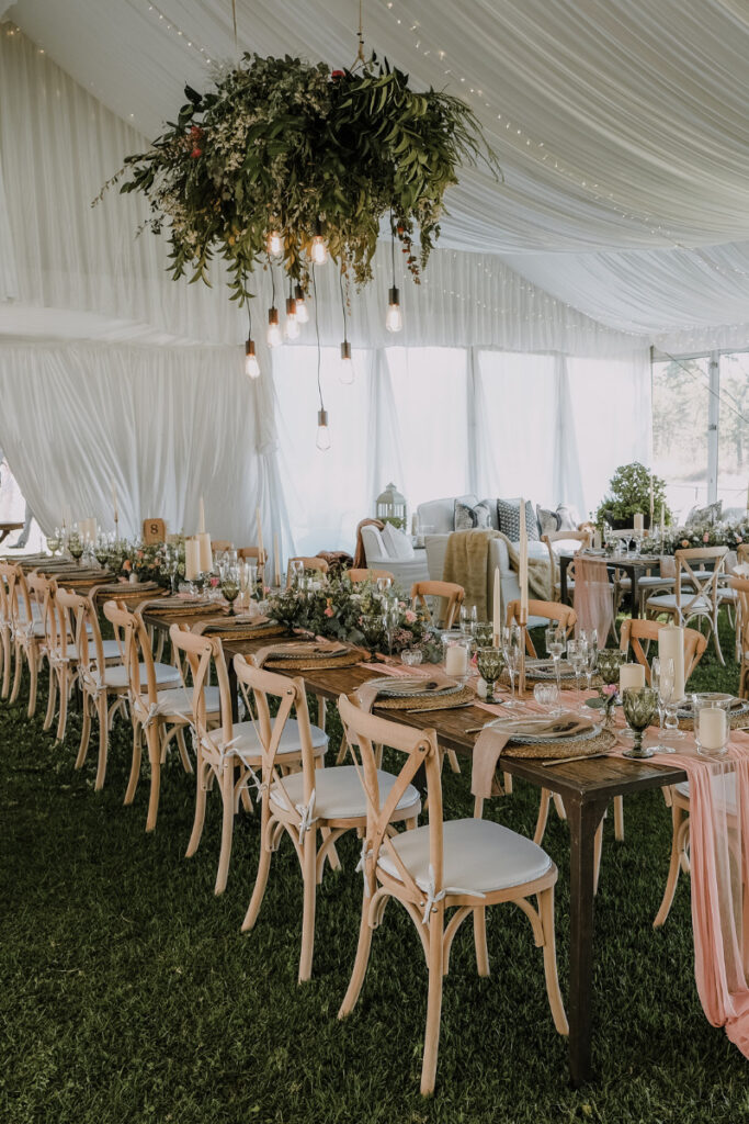 Elegant outdoor wedding reception setup under a white tent with chandeliers and round dining tables.