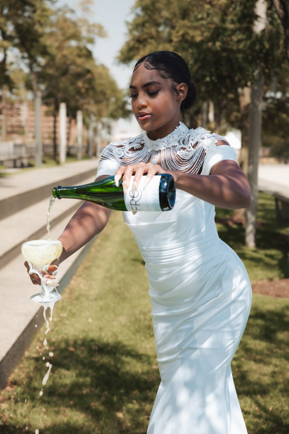 Bride in a white lace gown pouring champagne outdoors from a bottle into a glass tower.