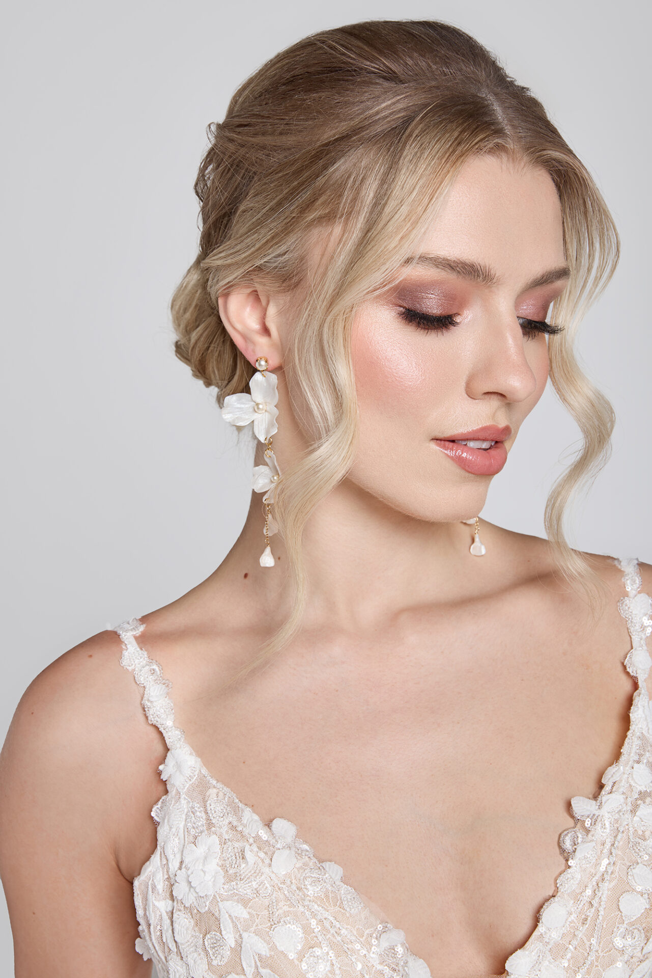 Bride with soft updo and pearl earrings, wearing a lace wedding gown, looking down with natural bridal makeup.