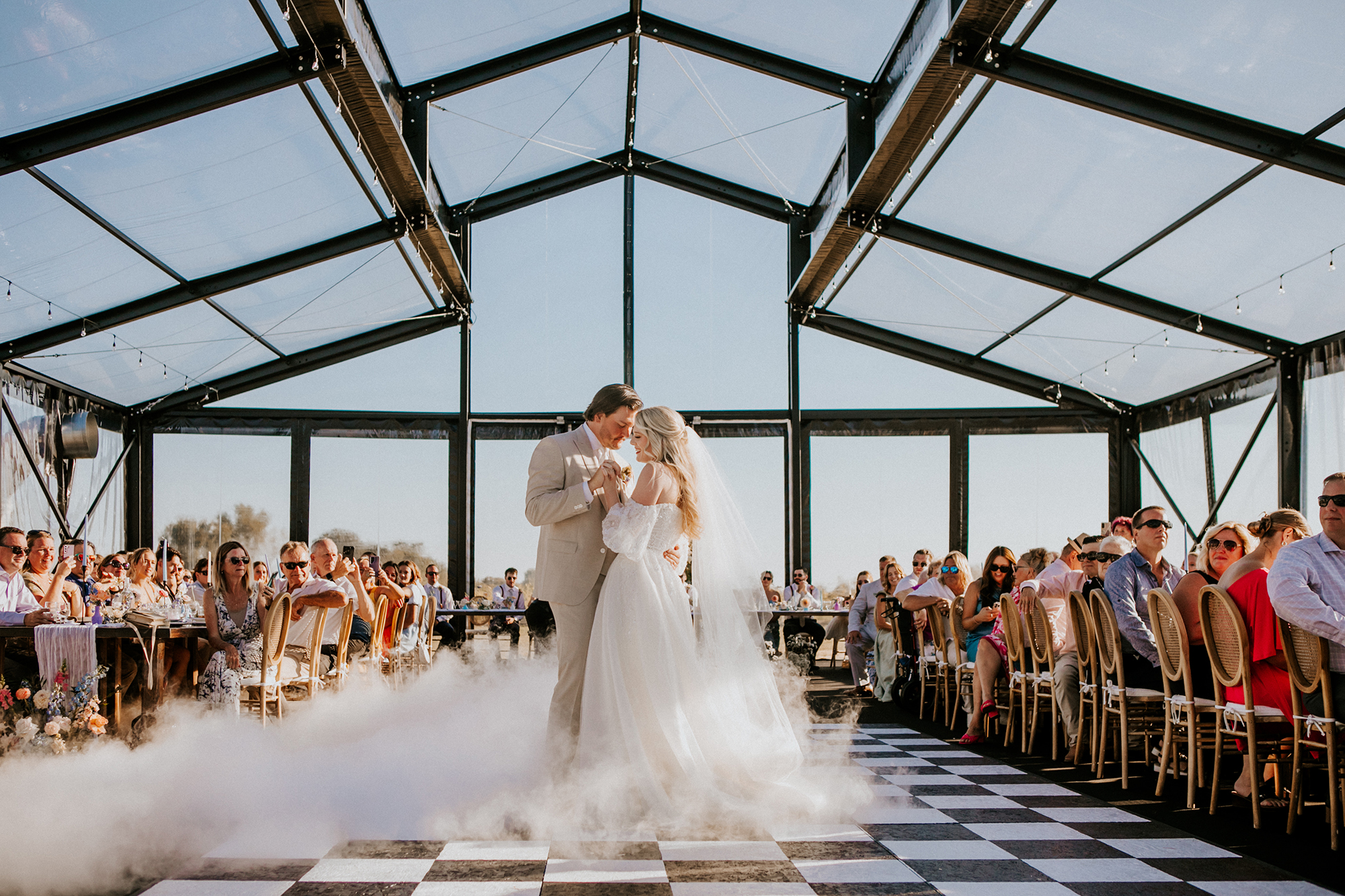 Bride and groom having their first dance on a checkered floor inside a glass-walled reception venue with guests watching.