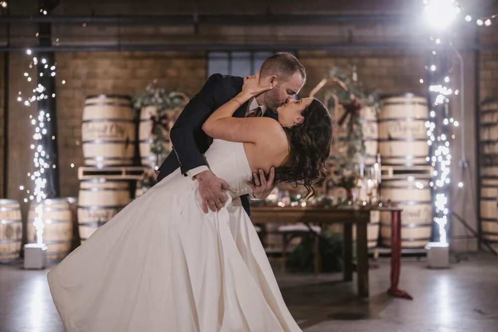 A groom dips the bride backward during a dramatic dance moment in a rustic venue decorated with barrels, twinkle lights, and winter greenery, while they share a kiss.