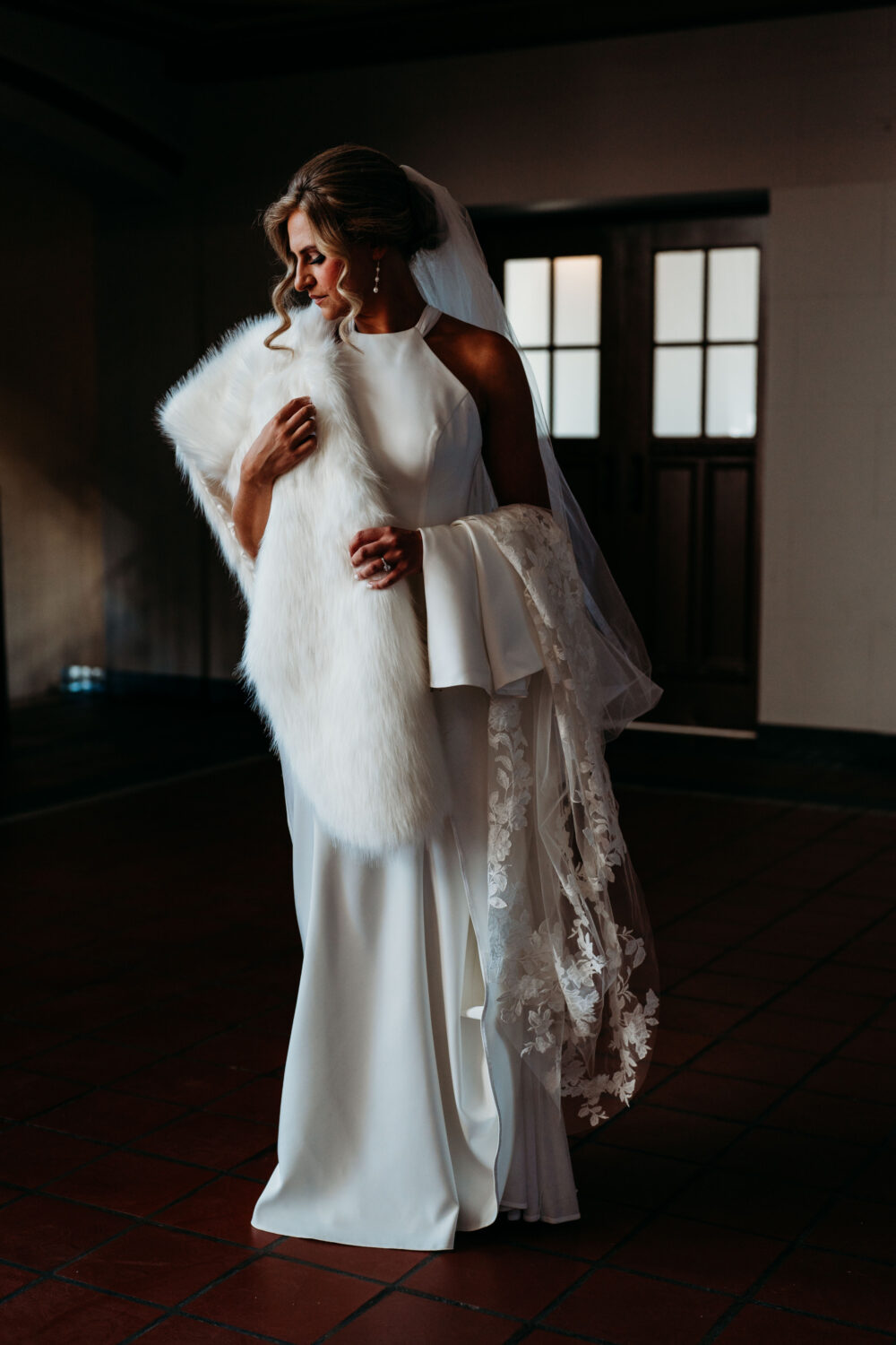A bride in a white halter-style wedding gown stands indoors holding a white faux fur stole and lace-trimmed veil, softly lit as she looks down with curled hair framing her face.