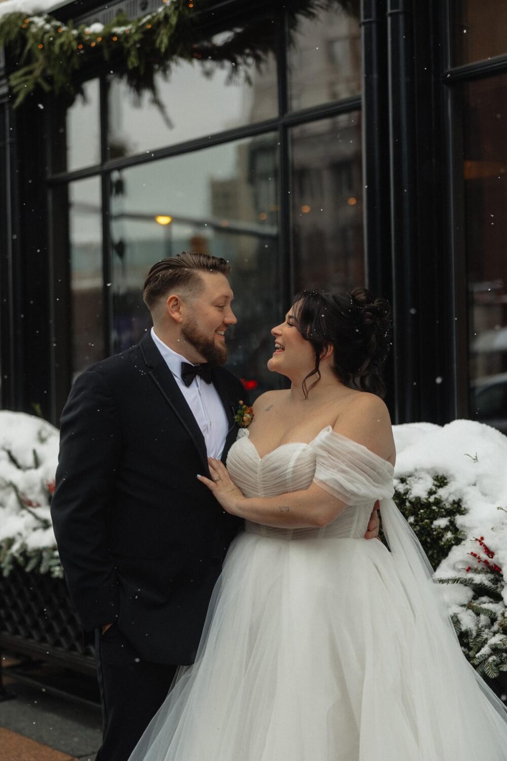 A bride and groom stand outside in the snow, smiling at each other; the groom wears a black suit, and the bride wears an off-the-shoulder tulle wedding gown surrounded by winter greenery.
