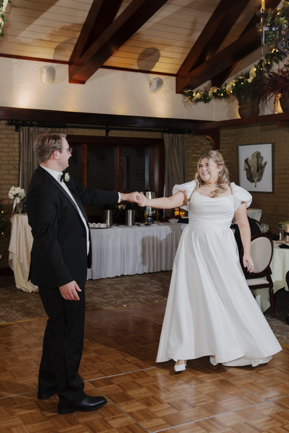 A bride and groom stand outside in the snow, smiling at each other; the groom wears a black suit, and the bride wears an off-the-shoulder tulle wedding gown surrounded by winter greenery.