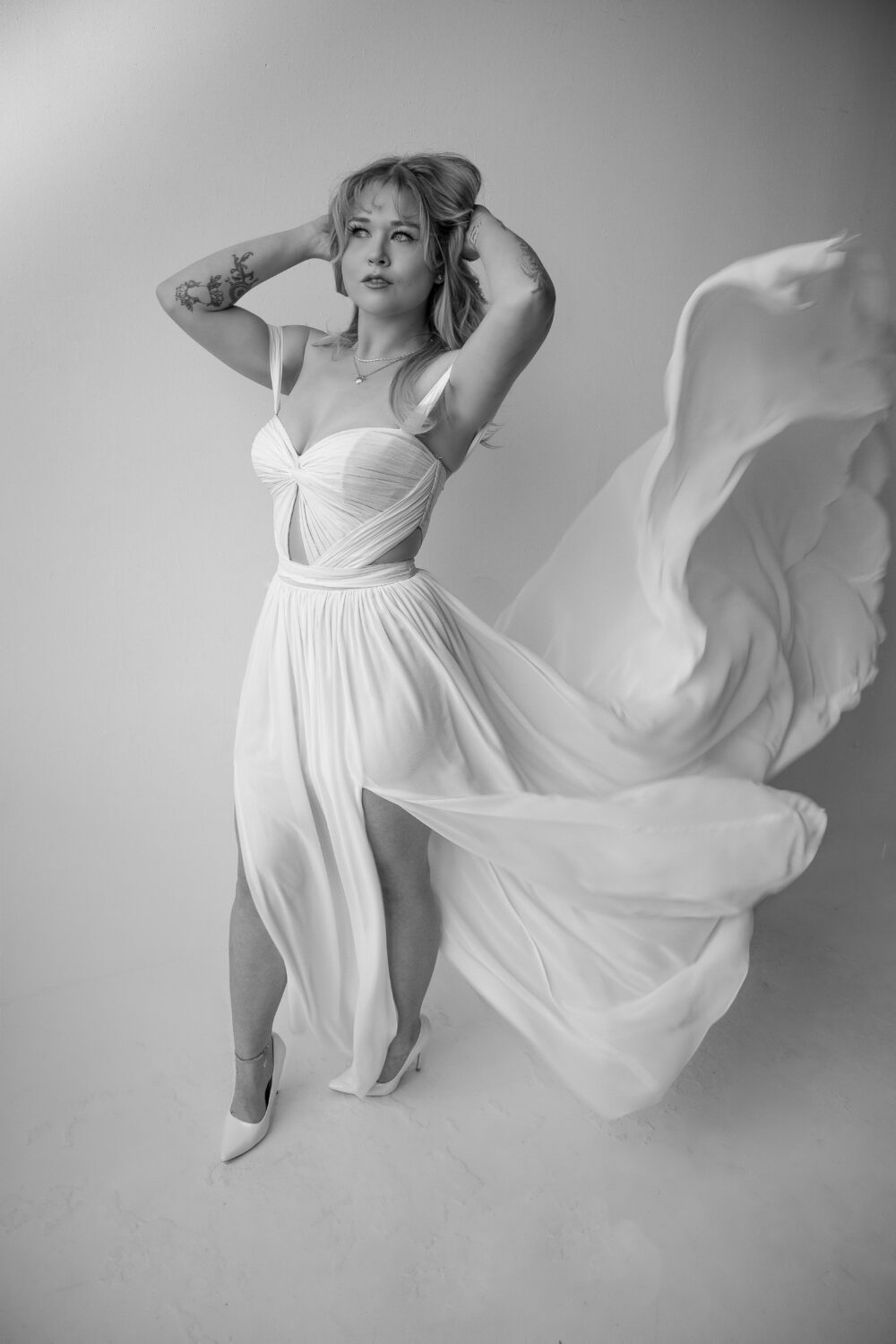 A bride wearing a flowy gown poses with her hands in her hair as the dress fabric sweeps dramatically to the side.