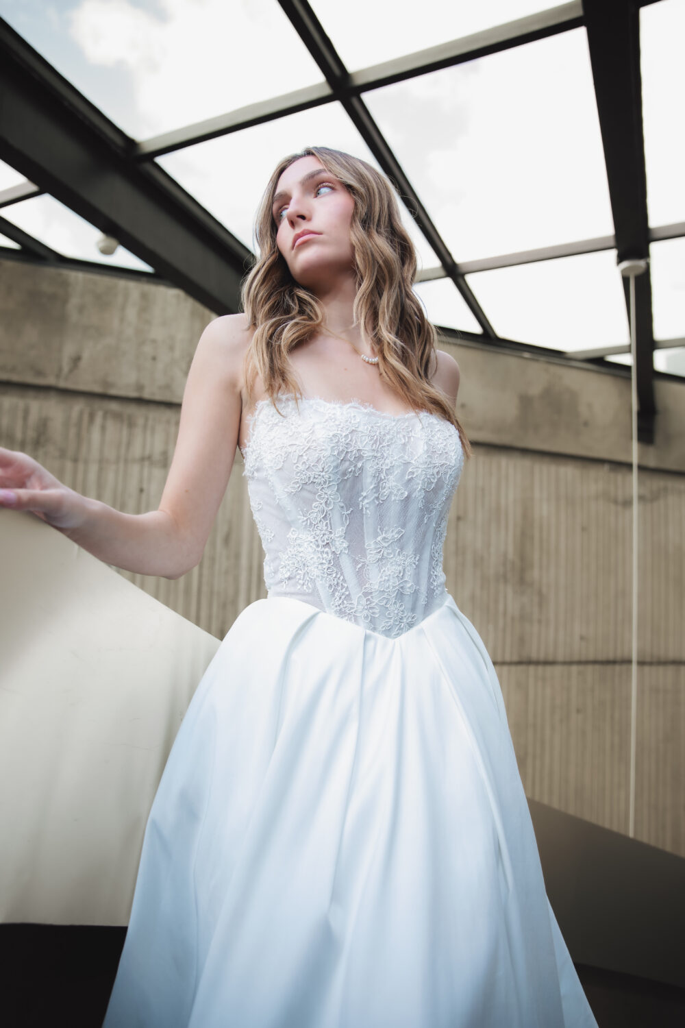A bride in a strapless lace-bodice ball gown stands under a glass ceiling, looking off to the side with one hand resting on a railing.