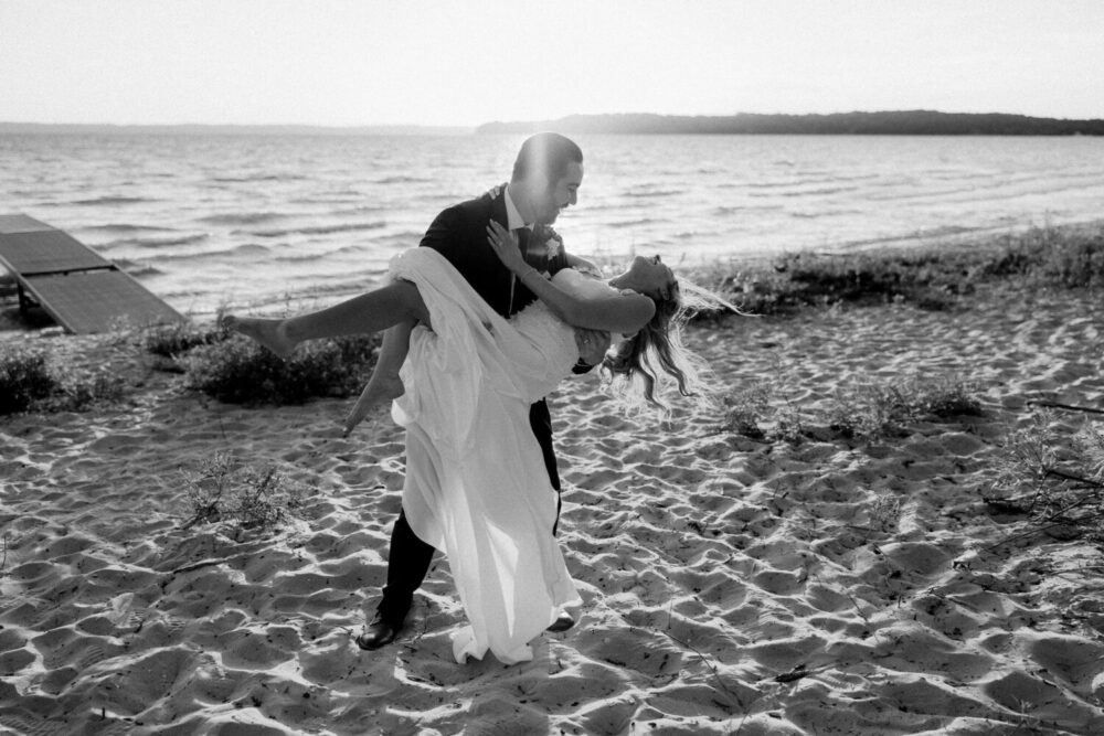 Black-and-white photo of a groom lifting his bride in his arms on a sandy beach, with waves and shoreline visible in the background.