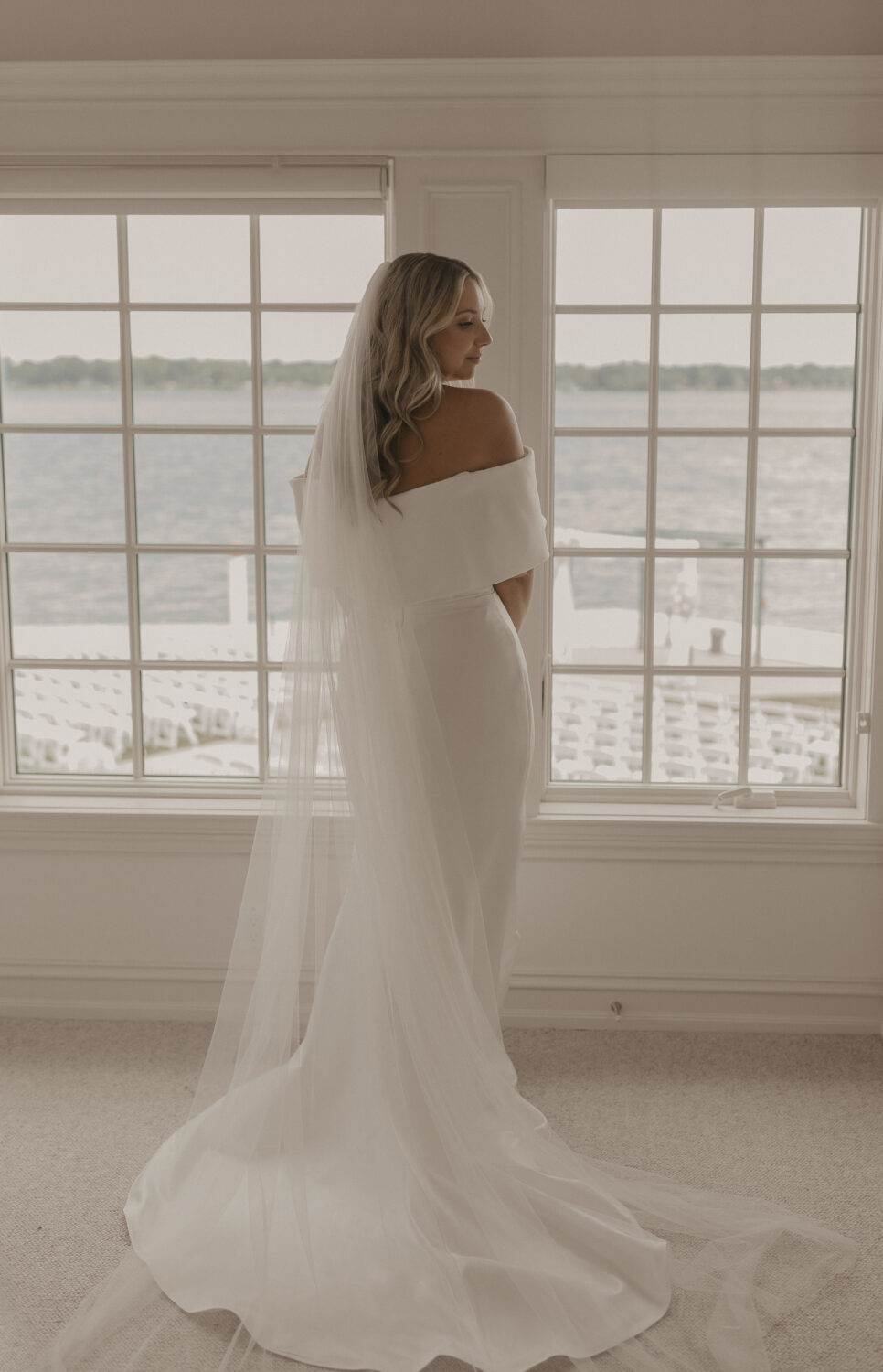 Bride standing by large windows in a minimalist room, wearing a fitted wedding gown with a long train.
