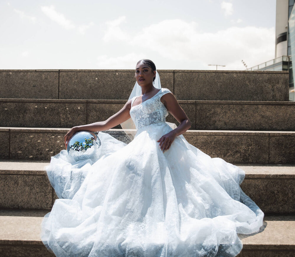 Bride in a white wedding dress sitting on stone steps, holding her gown and a disco ball.