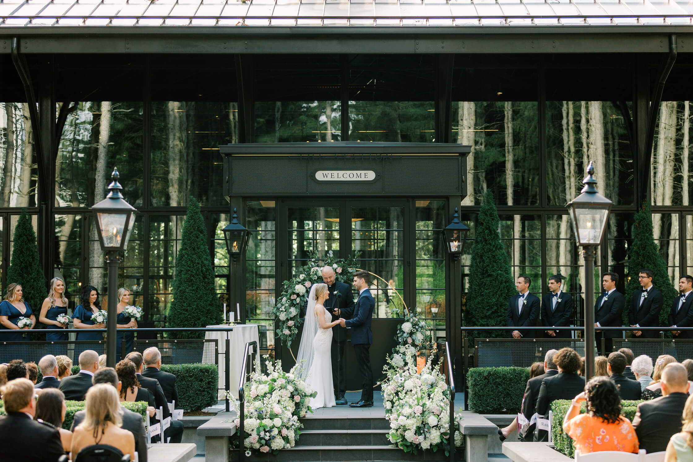 Outdoor wedding ceremony with bride and groom exchanging vows under a glass venue surrounded by greenery.
