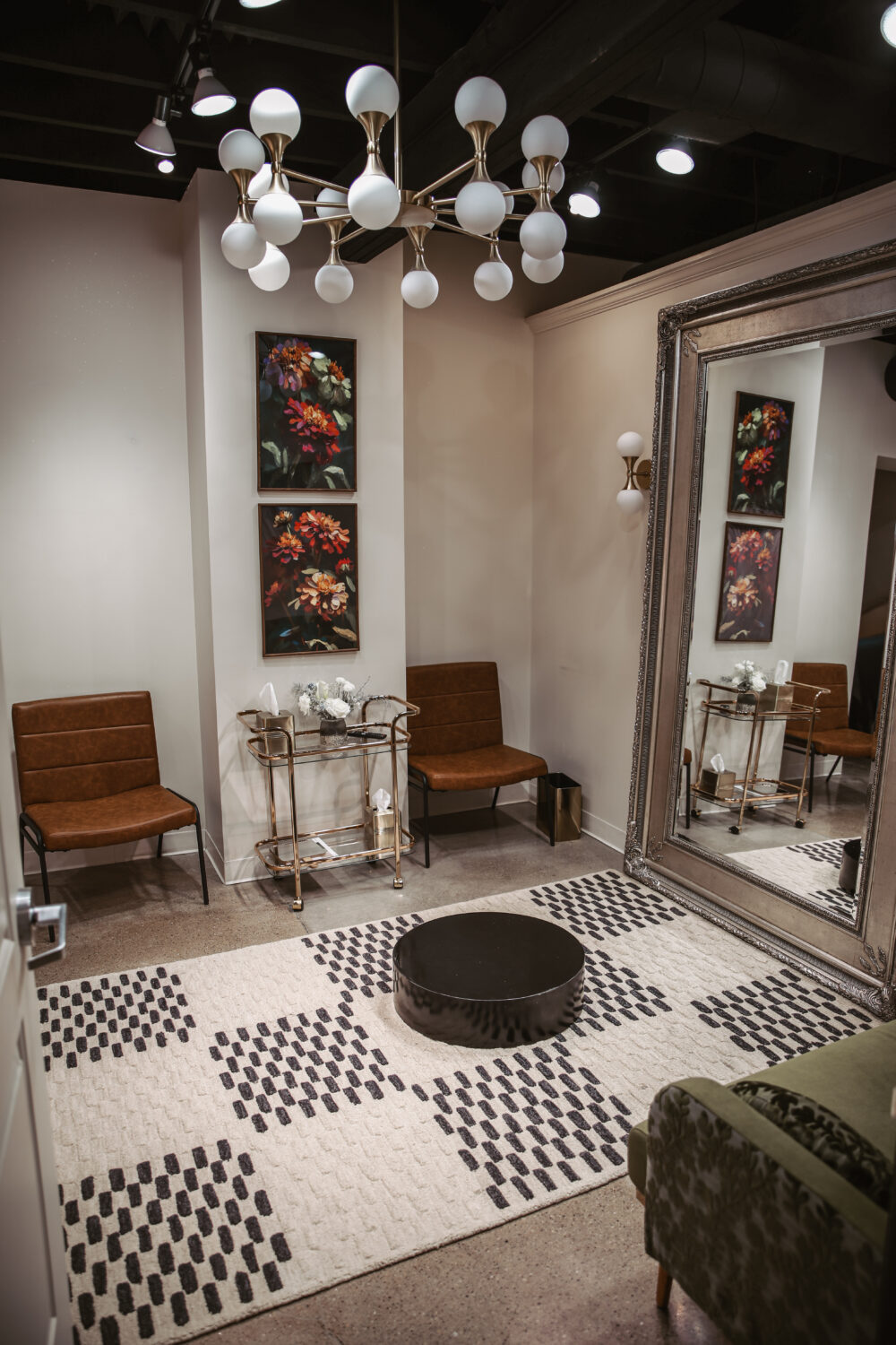 A private bridal fitting room with two brown chairs, floral artwork, a large mirror, and a patterned area rug on the floor.
