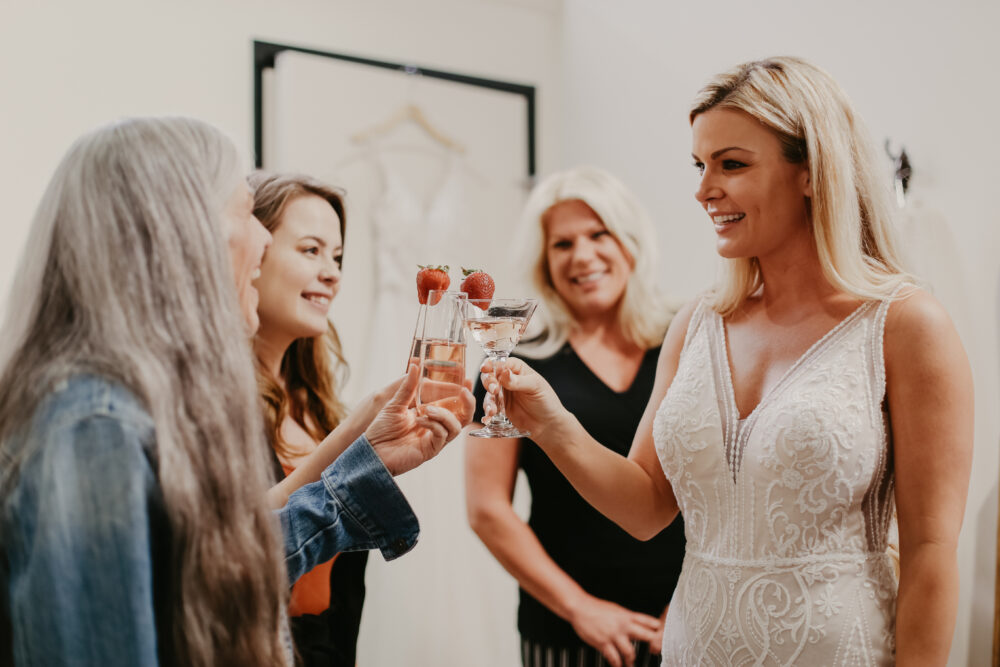 A bride in a lace gown clinks glasses with her family and friends, each holding drinks garnished with strawberries.