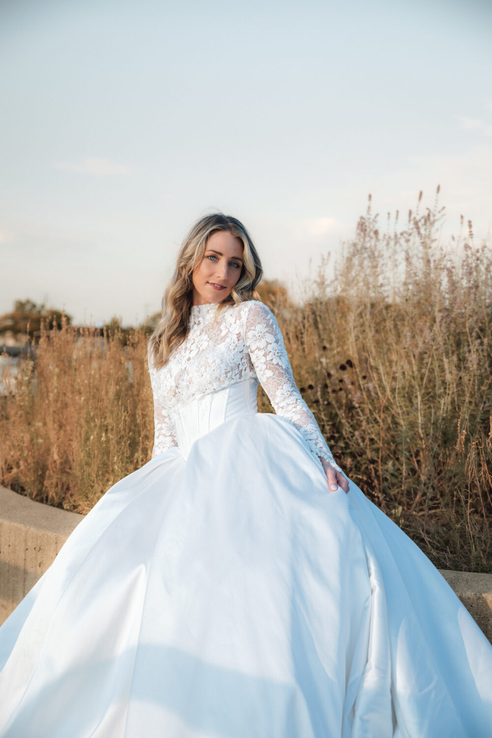 A bride wearing a long-sleeve lace bodice and full satin skirt stands outdoors near tall dried grasses, looking softly toward the camera.
