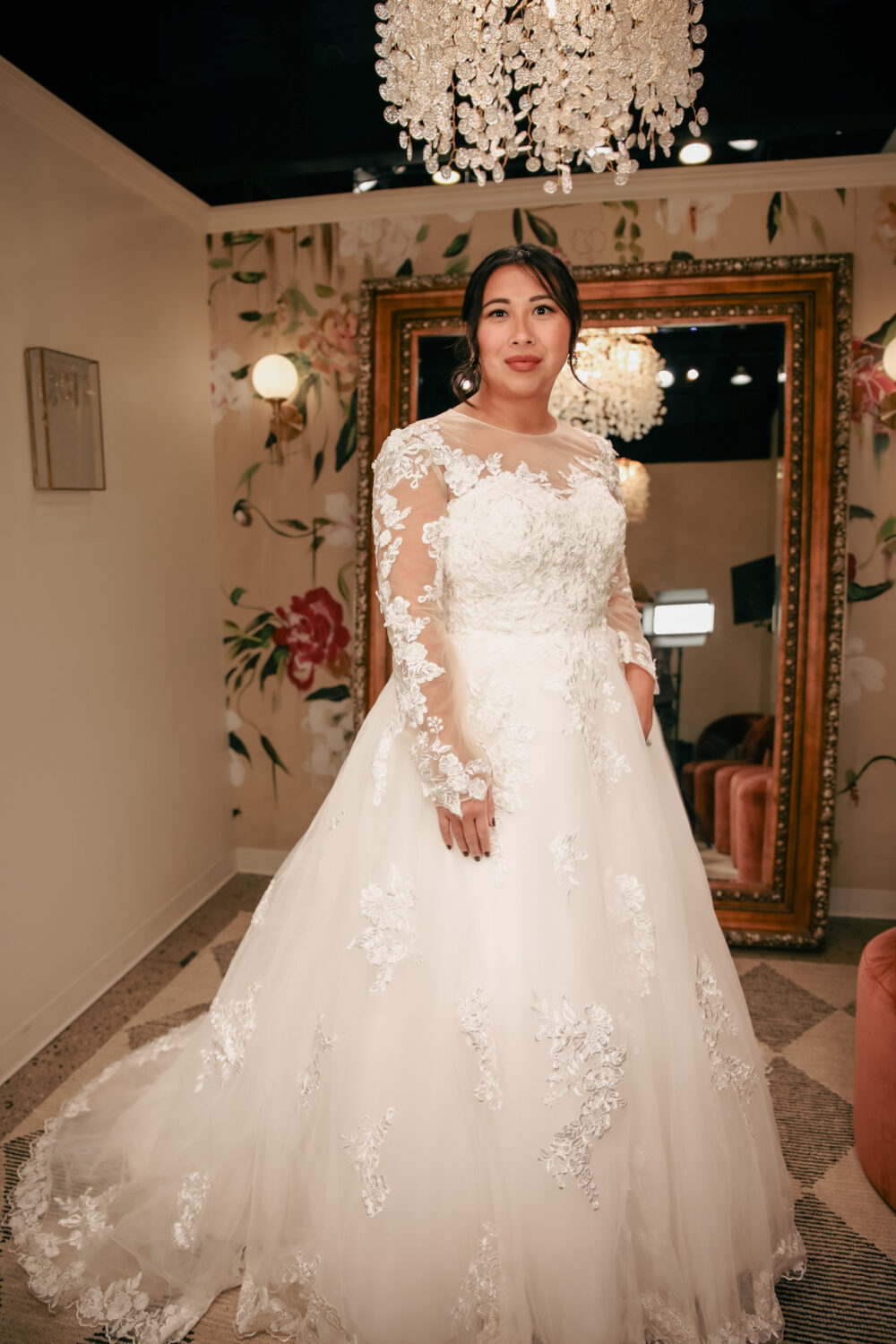 A bride stands in a floral-decorated fitting room wearing a long-sleeve lace wedding gown with an illusion neckline and a full tulle skirt.