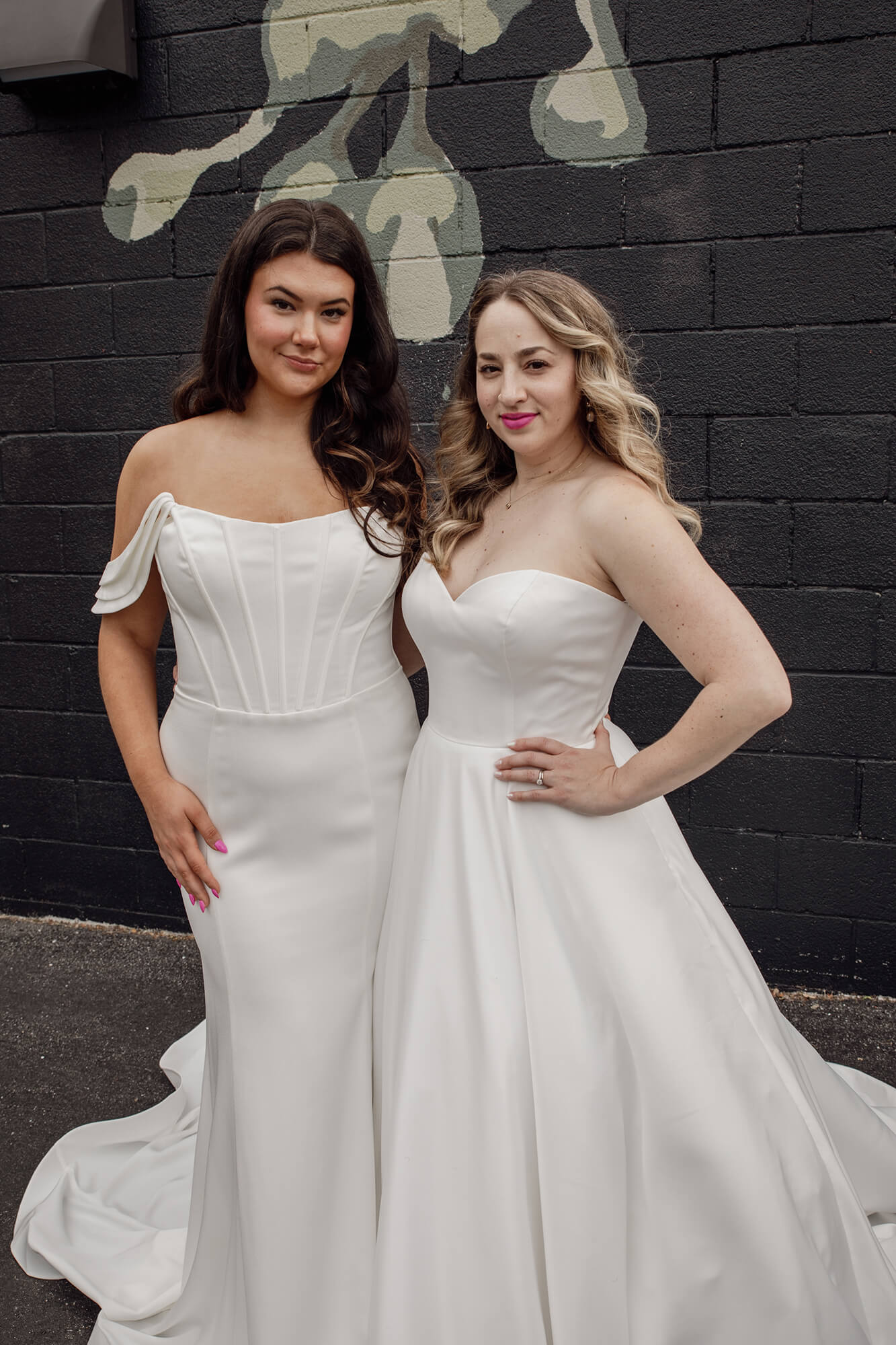 Two brides in off-shoulder white wedding dresses standing against a brick wall and smiling.