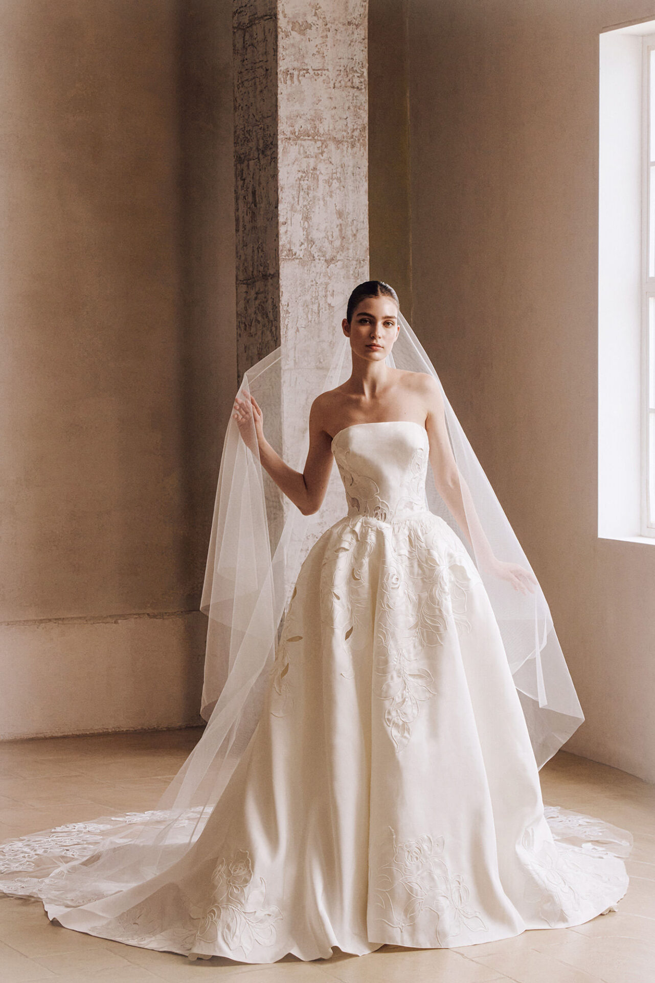 Bride in a classic strapless ball gown wedding dress standing beside a large window with soft natural light.