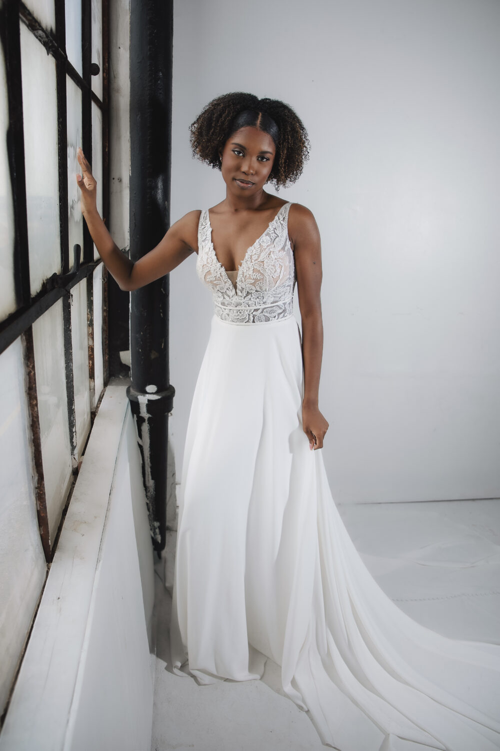 A bride wearing a lace bodice and chiffon skirt stands by a large industrial window, resting one hand on the frame.