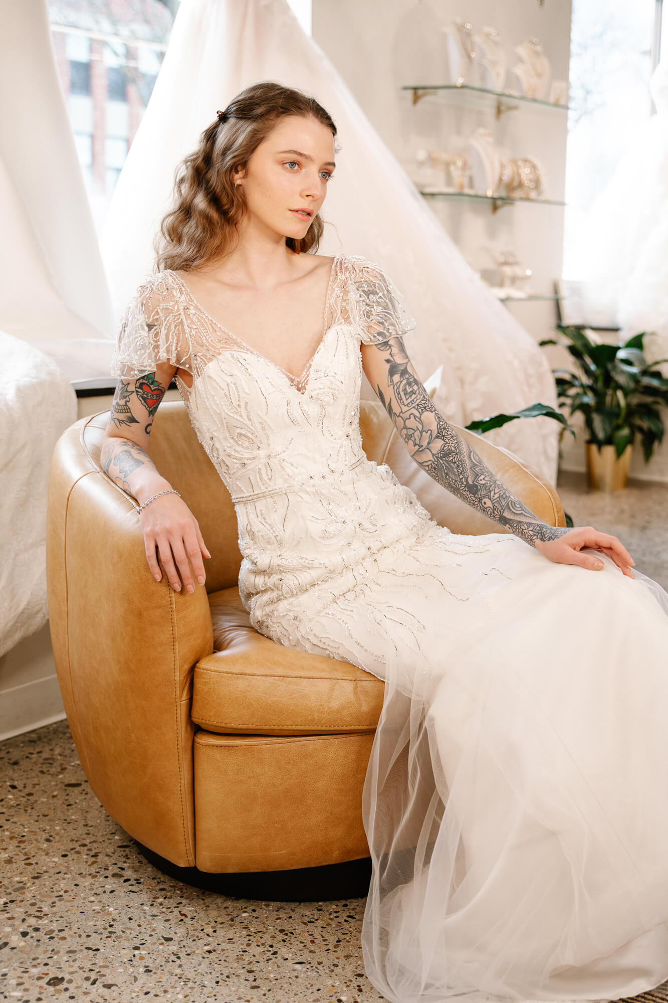 Bride seated on a tan chair wearing a lace-sleeved wedding dress with an illusion neckline and embroidered details.