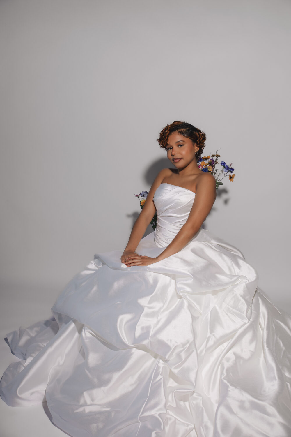 A bride in a strapless satin ball gown poses seated with her hands resting in her lap and flowers behind her.