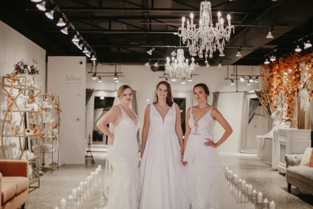 Three brides posing together inside a bridal boutique, each wearing a different white wedding gown.