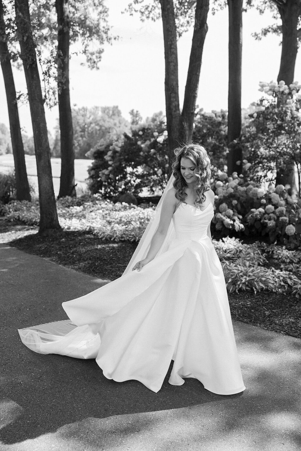 Bride outdoors in a wooded area, twirling in a flowing wedding dress, photographed in black and white.