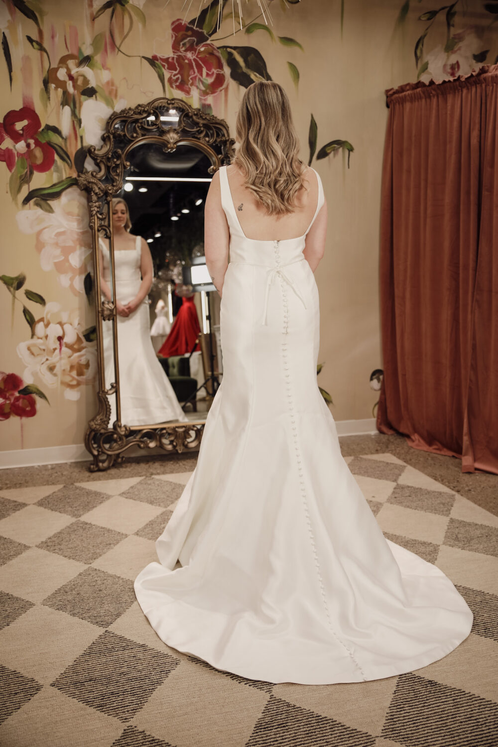 A bride wearing a fitted wedding dress stands with her back to the camera, looking at her reflection in a decorative full-length mirror.