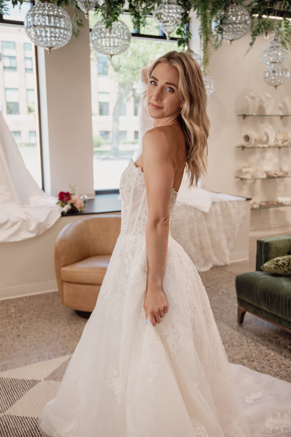 A bride in a lace wedding gown turns slightly toward the camera inside a bridal boutique with chandeliers and display shelves in the background.