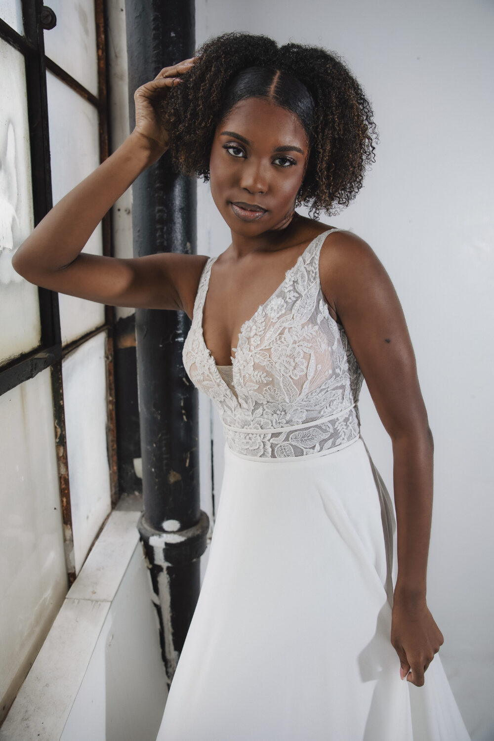 A bride with natural curls poses beside a tall industrial window while wearing a lace bodice and flowing skirt.