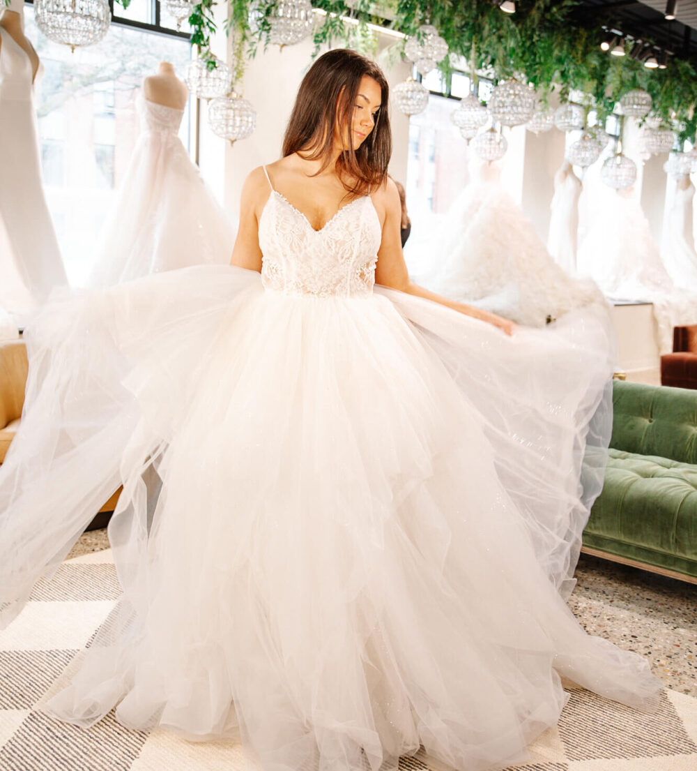 Bride wearing a voluminous tulle wedding gown twirling in a bridal shop with dresses on display behind her.