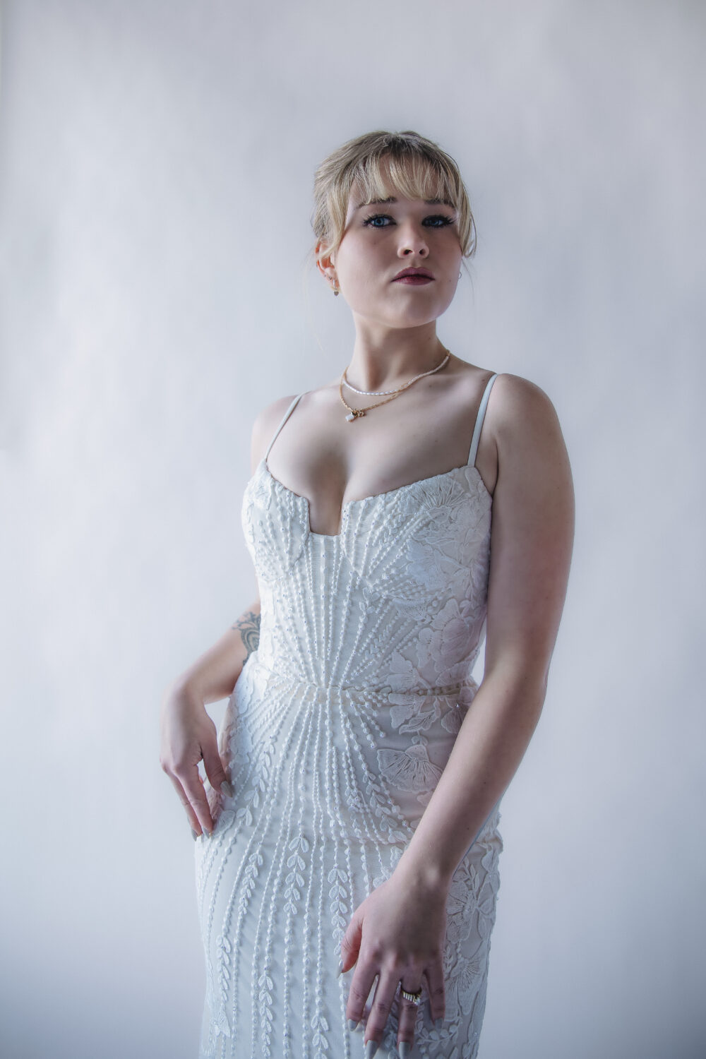 A bride wearing a fitted lace gown with thin straps poses against a soft, bright background.