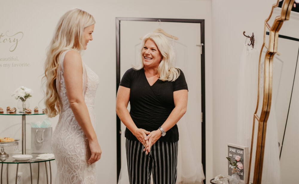 A bridal stylist smiles while speaking with a bride who is wearing a fitted lace wedding gown during a dress appointment.