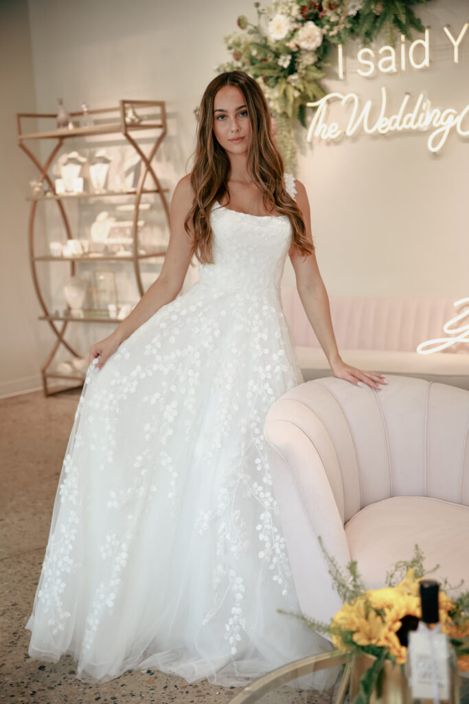 Bride sitting on a pink sofa wearing an off-the-shoulder lace A-line gown with floral appliqués.