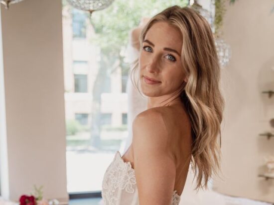 A close-up of a bride wearing a lace wedding gown looks over her shoulder inside a softly lit bridal showroom.