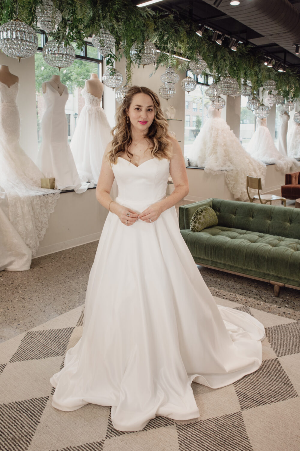 A bride in a sweetheart-neckline a-line wedding dress stands inside a bridal boutique with mannequins and chandeliers in the background.